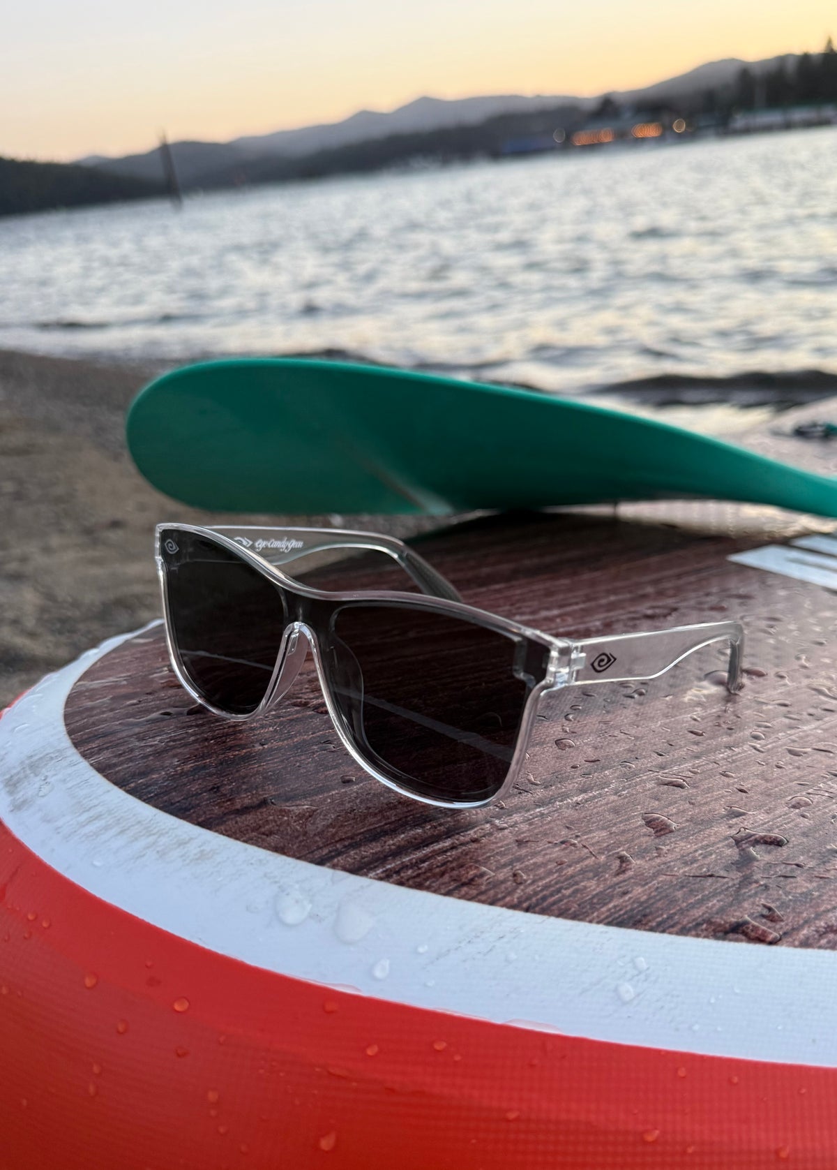 A pair of square sunglasses with clear frames and dark lenses resting on a paddleboard outdoors by the water, with a paddle and shoreline visible in the background.