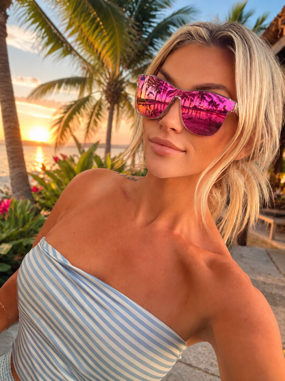 A woman wearing reflective pink cat-eye sunglasses with a palm tree sunset reflection, posing outdoors near palm trees and water at sunset.
